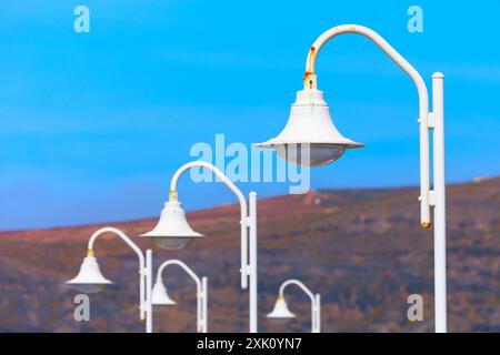 Row of white street lights illuminating in the evening. The lights are white and are hanging from poles Stock Photo