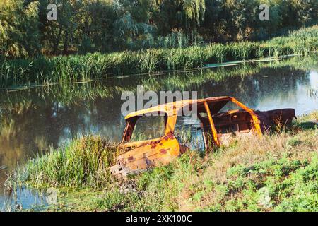 Rusted car is sitting in a swampy area near a river. Rusty car abandoned in the middle of the flowing river. Scene is eerie and unsettling Stock Photo