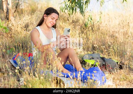 A young woman takes a break from hiking, comfortably seated among wild grasses, using her smartphone with a backpack beside her, under the shade Stock Photo