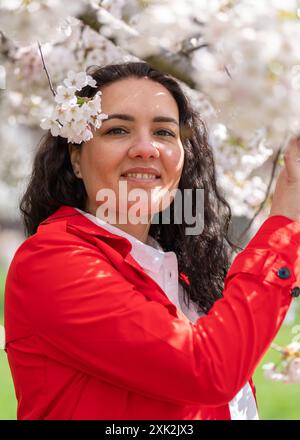 romantic image of a stylish woman in a red coat and white blouse. Positive mood. A cute girl gently holds a white sakura branch and looks at the camer Stock Photo