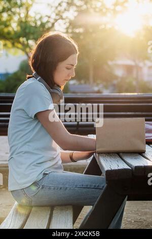 Side view of a young woman concentrates on writing in her notebook while sitting on a park bench and table, with headphones around her neck and a sere Stock Photo