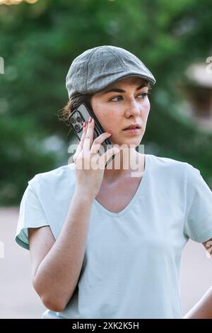 A focused young woman wearing a gray cap and a light blue t-shirt speaks on a smartphone, looking slightly away from the camera. Stock Photo