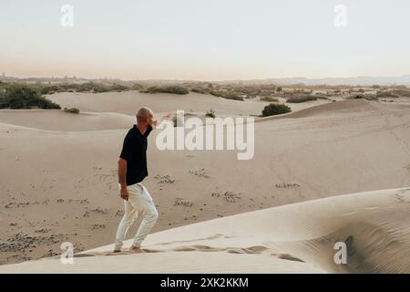 A bald man in a black shirt and white pants points into the distance while walking along serene sand dunes, under a soft summer sky, embodying a momen Stock Photo