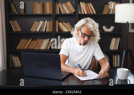 Middle aged senior woman using laptop computer writing notes at workplace. Focused mature old businesswoman work at office making written records doing online work. Mature boss professional worker Stock Photo