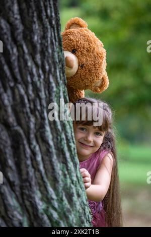 A sweet, pretty girl of 6 years old with long hair laughs cheerfully and looks out from behind a large tree. A large toy teddy bear peeks above her Stock Photo