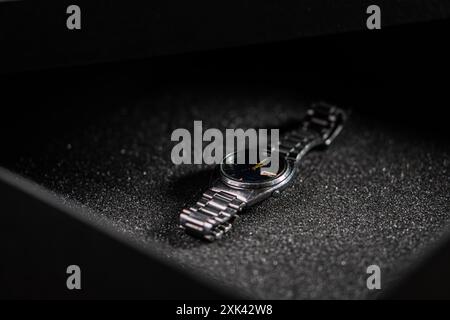 A sleek black wristwatch resting on a textured black surface inside a box. The watch features a minimalist design with a dark dial and metallic band, Stock Photo