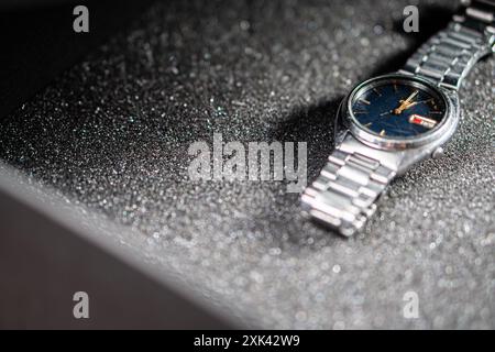 A close-up shot of a silver wristwatch with a dark blue dial, placed on a textured black surface. The watch features a date display and a metallic bra Stock Photo