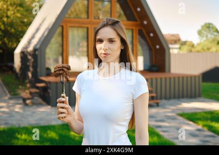 Cute young woman standing at backyard of a summer house and holds a barbecue fork with slices of steak in her hand Stock Photo