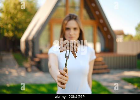 Defocused young woman stretching forward a barbecue fork with slices of steak in her hand Stock Photo
