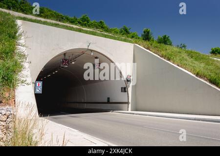 A concrete tunnel entrance with traffic lights and a paved road. The tunnel is framed by grassy hillsides on either side. Stock Photo