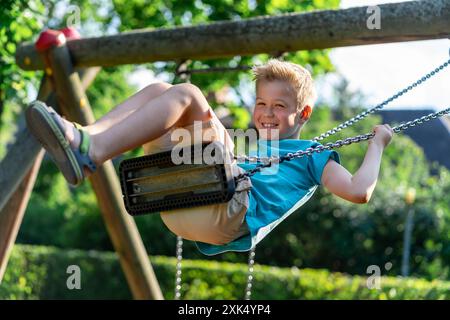 A boy in a blue T-shirt rides on a swing and smiles. Stock Photo