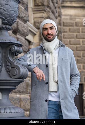 A smiling young Asian man in a light coat and a white knitted hat is traveling around the world. Taking in the sights of a new place. Close-up portrai Stock Photo