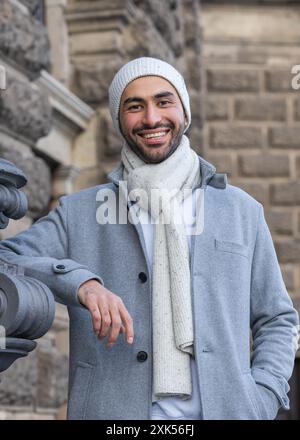 A smiling young Asian man in a light coat and a white knitted hat is traveling around the world. Taking in the sights of a new place. Close-up portrai Stock Photo