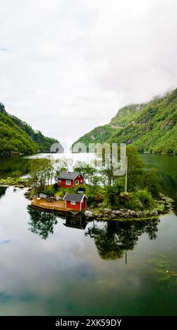 A small island with two red cabins nestled among green trees sits in a fjord, with mountains rising in the distance. Lovrafjorden, Norway Stock Photo