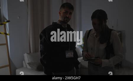 A man and woman, likely detectives, scrutinize evidence at an indoor crime scene, with one holding a notebook. Stock Photo
