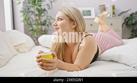 A pensive young woman lying on her bed holding a mug, looking serene in a cozy bedroom setting. Stock Photo