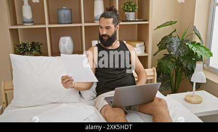 Handsome bearded man reviewing documents on laptop in cozy bedroom interior. Stock Photo