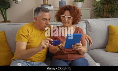 Middle-aged couple sitting together on a couch at home using a tablet and credit card, showing love and teamwork in a modern indoor living room settin Stock Photo