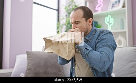 A bald man with a beard in a denim shirt breathes into a paper bag while sitting on a sofa indoors to calm an anxiety attack. Stock Photo