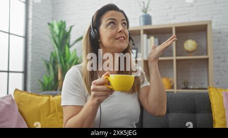 An attractive young woman wears headphones and holds a yellow cup, expressing a cheerful sentiment indoors. Stock Photo