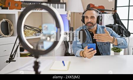 Hispanic senior man with grey beard in a music studio making video call indoors while wearing headphones. Stock Photo