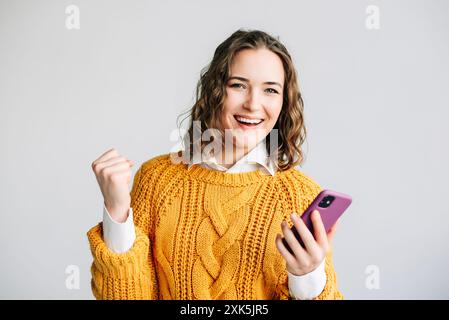 Excited Woman Celebrating with Smartphone. A joyful young woman celebrating good news on her smartphone. Standing against a white background, she Stock Photo