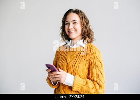 Happy Woman Using Smartphone. Smiling young woman with curly hair using her smartphone. She is browsing the internet, standing against a white Stock Photo