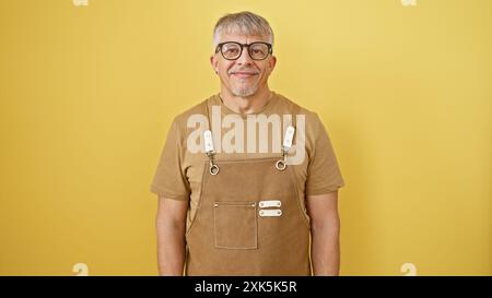 A smiling grey-haired man wearing glasses and overalls posing against a yellow isolated background. Stock Photo