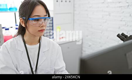 A young asian woman wearing safety glasses and a lab coat working in a laboratory environment, focused on her research. Stock Photo
