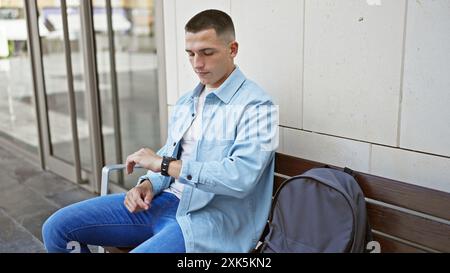 A young hispanic man checks time while seated on a bench outdoors in an urban city. Stock Photo