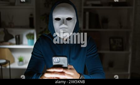 Masked person in hoodie using smartphone indoors, evoking thoughts of privacy, technology, and mystery. Stock Photo