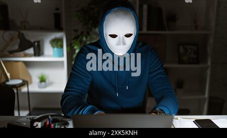 Mysterious person with a white mask and hoodie using computer in a dark office setting at night. Stock Photo