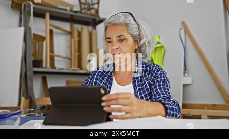 A focused mature woman carpenter using tablet in a well-lit workshop with woodworking tools. Stock Photo