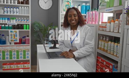 Woman working in a pharmacy, smiling while using a laptop, surrounded by shelves of pharmaceutical products and medical supplies. Stock Photo