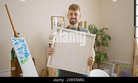 Handsome man with beard holding a blank canvas in a bright art studio. Stock Photo