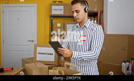 A young man wearing a headset and badge marks inventory on a tablet in a donation center warehouse. Stock Photo