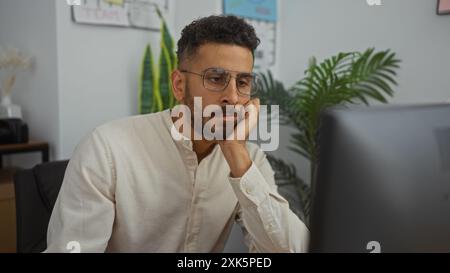 Handsome young hispanic man working in an office setting, wearing glasses and a casual shirt, focused on a computer screen. Stock Photo