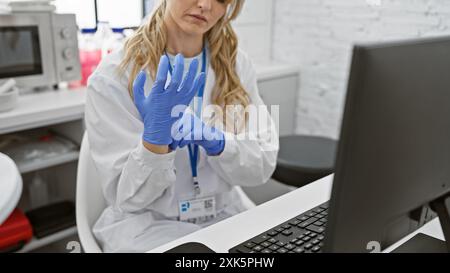 A focused young caucasian woman in a laboratory wearing a lab coat and blue gloves working with a computer. Stock Photo