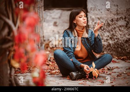 A young woman sits cross-legged in an alleyway, blowing on a leaf while surrounded by fallen foliage. Stock Photo