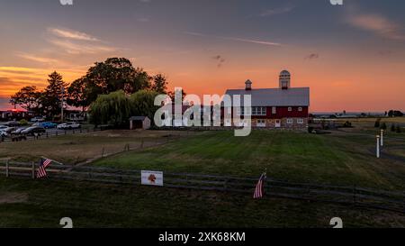 Ronks, Pennsylvania, USA, July 19 2024 - Large Red Barn With Silver Roof And Twin Cupolas At Sunset, Surrounded By Open Fields, A Weeping Willow, Fencing, And Other Farm Structures Under A Pink And Orange Sky. Stock Photo