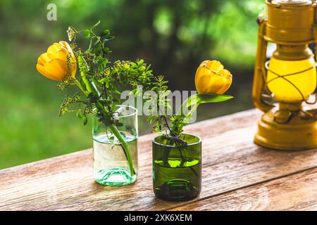Vases with yellow tulips on a wooden garden table, decoration made from green wine bottles, craft tip Stock Photo