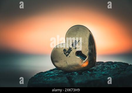 Old gold-colored lock in heart shape with the text love you on a wooden pole with the sunset on the beach in the background Stock Photo
