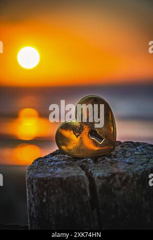 Old gold-colored lock in heart shape with the text love you on a wooden pole with the sunset on the beach in the background, vertical Stock Photo
