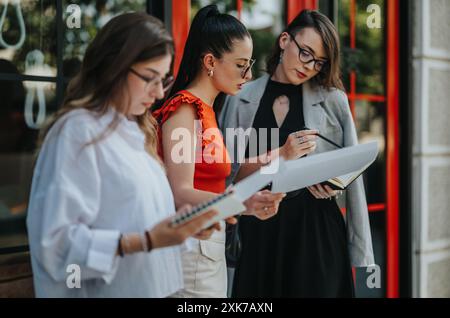 Businesswomen reviewing documents outside office building, discussing strategies and planning work. Professional women collaborating outdoors. Stock Photo