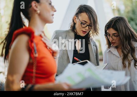 Three businesswomen collaborating outdoors, reviewing documents and discussing a project Stock Photo