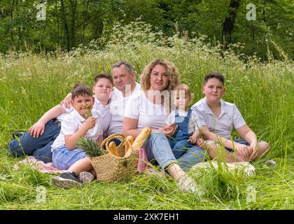 Portrait of a large, cheerful family at a picnic. They sit in the thick green grass. Happy dad and mom and four sons of different ages. The smallest c Stock Photo