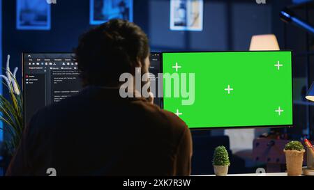 Programmer typing complex code on green screen computer in neon lit home office, developing software application. Teleworking IT specialist doing remote project tasks on mockup PC monitor, camera A Stock Photo