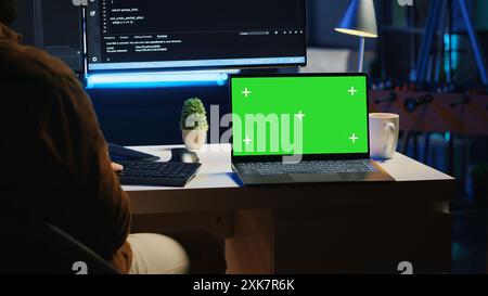 Green screen laptop next to freelancing man doing IT support job from apartment office, coding on PC. Mockup notebook on desk where software engineer writes lines of code on computer at home, camera A Stock Photo