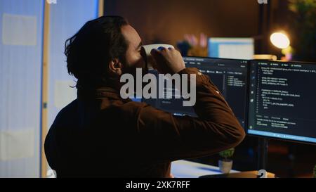 Computer scientist at home office desk writes binary code scripts and drinks from mug of coffee. IT expert in apartment enjoying tea while using programming to safeguard consumer data, camera A Stock Photo