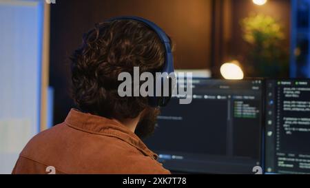 Man doing IT support job from apartment office, drinking cup of coffee and listening music while coding. Developer with headphones on enjoying hot tea and writing code on computer, camera A close up Stock Photo